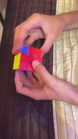 A pair of hands are shown holding a cube, which is being manipulated and spun. The cube is a Rubik's cube with colorful faces. The hands are turning the cube rapidly. The background is a textured surface that appears to be a bedspread.