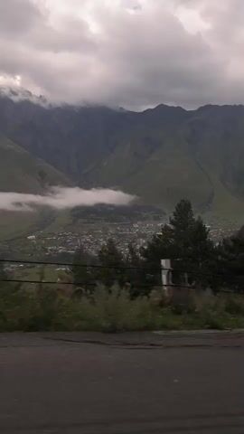 The video shows a view from a vehicle moving along a paved road. The road is dark asphalt, and the surrounding area is covered in green grass and some sparse vegetation. In the middle ground, there are several pine trees and a fence line with white posts. Beyond the trees, a valley unfolds with a small town or village nestled among the green hills. Majestic, rugged mountains dominate the background. The peaks of these mountains are shrouded in dramatic, grey clouds, with some sections of mist clinging to the lower slopes. The sky is overcast with heavy, dark clouds, suggesting a potentially stormy or overcast day. The perspective indicates the camera is likely positioned inside a car, looking out a window or windshield. The movement of the vehicle is evident through the slight blur and changing perspective of the scenery.
