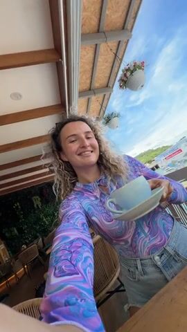 The video shows a woman with curly blonde hair and a bright purple and blue tie-dye top smiling and winking at the camera. She is holding a cup of green matcha latte with latte art. She is wearing denim shorts. The background shows a tropical outdoor setting with a balcony, chairs, plants, and a view of a body of water with boats and buildings. The woman is in focus, and the background is slightly blurred. The lighting suggests it is daytime.