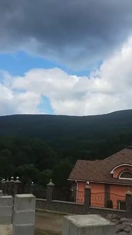 The video shows an exterior view of a house with an orange facade and a brown roof, partially obscured by a stone fence. In the background, there is a densely forested hill or mountain under a cloudy sky. The scene appears to be in a rural or suburban setting.