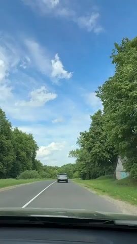 The video shows a view from inside a car driving on a road. There's a silver car ahead on the road, traveling in the same direction. The road is surrounded by green trees and grass. The sky is bright blue with scattered white clouds. A white line marks the center of the road.  The dashboard is visible at the bottom of the frame.  Music is playing.