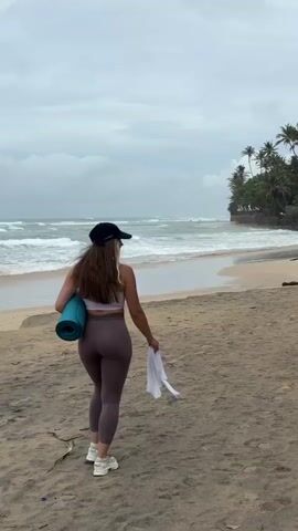 A woman walks along a sandy beach, carrying a rolled-up blue yoga mat in her left arm and a white towel in her right hand. She is wearing a light pink sports bra, dark purple leggings, white sneakers, and a dark baseball cap. The ocean with moderate waves is to her left, and a lush green cliffside with palm trees is to her right. The sky is overcast. The camera is positioned behind the woman, capturing her from a medium distance as she walks away from the viewer and towards the cliff.