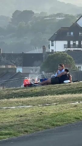 A medium shot captures a couple sitting and lying on a grassy hill, with a picturesque background of houses and trees enveloped in mist. The woman, wearing a light blue jacket and dark shorts, sits up and appears to be talking or gesturing. The man, wearing a dark tank top and shorts, lies on his back. A bag is placed behind them, and a red bucket sits to their left. Smoke or steam rises in the background behind a fence. The environment consists of green grass in the foreground, with houses and trees in the hazy, distant background. The lighting is soft, suggesting either early morning or late afternoon.