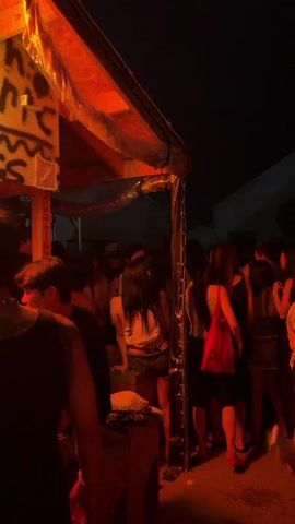 The video shows a dimly lit outdoor gathering at night. A group of people are visible, some standing and talking, others appearing to be in motion. The lighting is predominantly red and orange, creating a warm, somewhat hazy atmosphere. In the foreground, a person's back is visible, with part of their arm showing. Behind them, a makeshift structure with a transparent covering is present. The background reveals more individuals and hints of surrounding buildings or structures. The overall impression is that of a casual, informal social event.