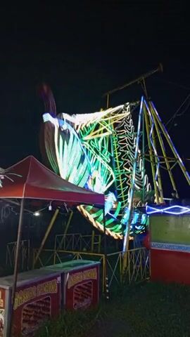 The video shows a carnival ride at night. The ride is lit with bright lights and is moving. There are food stands next to the ride.  The sky is dark and there are some trees in the background. There are no people in the video.