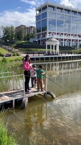 A medium shot shows a woman and two young boys standing on a wooden dock beside a pond. The woman, wearing a pink long-sleeved shirt and black pants, stands to the left. One boy, in a white shirt and gray pants, stands to her right, and another boy, in a green shirt and brown pants, stands on the far right, holding a fishing rod in the pond. Circular ripples are visible on the water's surface. In the background, a large building with a reflective glass facade and white arches is seen across the pond, with its reflection visible in the water. A white gazebo is also visible near the building. The sky is blue with some clouds.