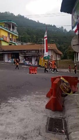 A group of children in orange shirts and black pants are walking across a street in a town. The street is paved and has orange traffic barriers. There is a tall flag with red and white stripes in the background, along with buildings and trees on a hillside. The sky is overcast. One adult male in a red shirt is also crossing the street with the children.