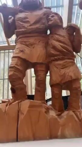 The video shows a large wooden sculpture of two figures, presumably men. The larger figure stands upright with a thick beard, holding a tool or log over his shoulder. The smaller figure stands beside him, looking up. The sculpture is situated in what appears to be a building with a glass ceiling and natural light filtering through. The carving style is rough, emphasizing the texture of the wood. There are informational plaques visible behind the sculpture. The camera pans slightly upward, focusing on the details of the sculpture.
