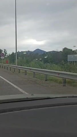 A car is driving on a highway on a cloudy day. The road has multiple lanes and a metal guardrail runs along the side. There is vegetation and trees on the right side of the road, and a distant mountain can be seen. A traffic sign is visible on the left side of the road. Other cars and a truck are also on the highway.