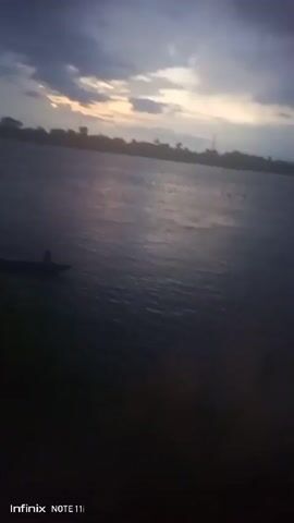 The video shows a still shot of a lake or river at dusk. A small boat with two people is visible in the water. The sky is cloudy with a hint of sunlight breaking through. The foreground is dark, with some greenery visible along the shore. The overall scene is calm and peaceful.  The video was shot with an Infinix Note 11i phone.