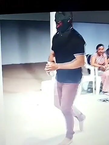 A man with a black mask covering his face and a black shirt stands in a room with white chairs and a concrete floor. He is wearing purple pants and is barefoot. In the first frame, he is standing and holding a bottle. In the second frame, he bends down and touches the floor with his hands. In the third frame, he stands up and raises his hands. In the fourth frame, another person in a red shirt and black pants stands in front of him. The man in the red shirt is also wearing a black mask. There are other people sitting in the chairs. The room appears to be outdoors or in a covered outdoor space.