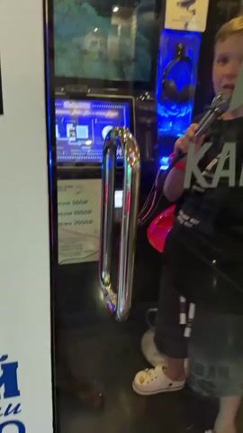 The video shows a young boy singing into a microphone inside what appears to be a karaoke booth. He is wearing a black t-shirt and shorts, with a red wristband on his left arm. He has blond hair and is looking towards the camera. The booth has a large screen displaying song options, headphones hanging on the wall, and a keypad for selecting songs. A metal handle is visible on the door of the booth. The boy's facial expressions suggest he is actively engaged in singing. The overall environment is dimly lit with blue and pink accents from the lighting inside the booth.