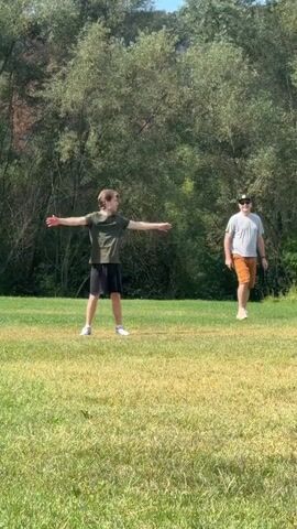 Two men are standing on a grassy field, with trees and a hill in the background. The man on the left is wearing a dark green t-shirt and black shorts, with white sneakers. He has his arms outstretched. The man on the right is wearing a grey t-shirt, orange shorts, a baseball cap, and sunglasses. He also has his arms outstretched. The man on the left moves his arms down, and the man on the right moves his arms down and then up. A volleyball appears in the air between the two men. Another man, wearing a black t-shirt and black shorts, runs into the frame from the right and lunges forward, reaching for the volleyball.