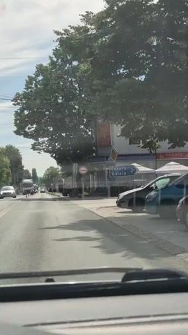 The video appears to be shot from the perspective of a driver. The scene depicts a street in a town or village. On the right side of the road, there is a white building with a tiled roof. Several cars are parked in front of it, and there is a sign that says "Galata". Further down the street, more cars and some trees are visible. On the left side of the road, there are more buildings, trees, and some people walking. There is also a truck on the road. The sky is cloudy and the lighting suggests it is daytime. A traffic sign with a red circle and a diagonal line is visible on the left. Another sign with a yellow diamond shape is also visible near the building on the right. The overall impression is of a quiet, possibly rural or suburban street.