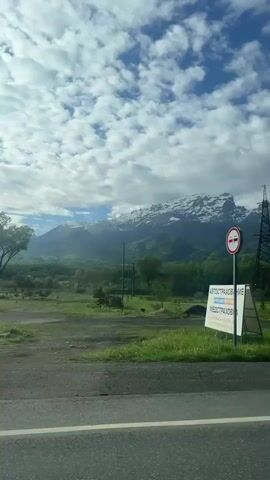 A vehicle passes an electrical station beside a road, with snow capped mountains in the background. The sky is blue with many clouds. Power lines and poles are plentiful throughout the environment.