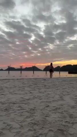 The scene takes place on a sandy beach under a dramatic sky painted with hues of pink, orange, and grey at what appears to be dawn or dusk. The sky is filled with textured clouds. In the distance, dark silhouettes of islands punctuate the horizon above the water line. A line of tall, dark structures resembling basketball hoops is visible in the shallow water near the shore, adding a geometric element to the landscape.

Two figures are present on the beach. Closer to the camera, a person in dark swimwear is walking in the sand. Further away, near the water's edge, a smaller figure, possibly a child, is running and playing. The grainy texture of the sand suggests a fine grain size. The overall tone of the scene is peaceful, highlighted by the soft colours of the sky and the silhouettes of the landscape.