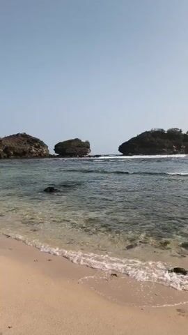 The video shows a beach with clear water and sand. There are a few people in the water and on the beach. There are also some rocks and trees in the background. The sky is blue and clear.