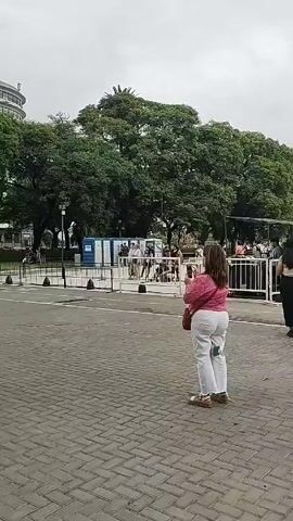 The video shows a small crowd gathering around a temporary stage set up in a paved public square. The stage is black and appears to be the back of a trailer, with a band setting up on it. The backdrop of the scene includes a building with a rounded corner and a large number of trees. A series of portable toilets are set up behind a metal barrier. Several people are standing and watching the band prepare, while others are taking photos. A sign on the side of the stage reads "Secretaría de Cultura." The overall atmosphere is casual and suggests an outdoor performance or event is about to begin. The sky is overcast.