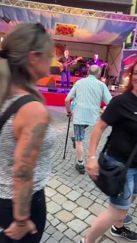 The video shows an outdoor concert with a band performing on a stage. The stage is decorated with a banner depicting a landscape with clouds and a picture of fruits. There are several people in front of the stage, some are dancing and others are watching. One person is using crutches to stand. The ground is paved with cobblestones. The lighting is colorful, with purple and pink lights illuminating the stage. There are speakers and musical equipment on and around the stage. The overall atmosphere appears to be festive and lively.