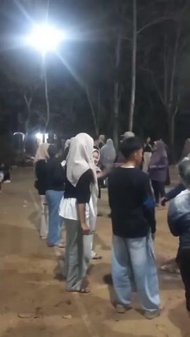 A group of people are gathered outdoors at night, illuminated by a bright overhead light and some flagpoles. The ground is dirt and some light-colored powder is visible on the ground and on the back of a person in the foreground. Many of the people are wearing hijabs and casual clothing. Some people are interacting, and appear to be engaged in a celebration or event. A banner in the background reads "Gelar Kemerdekaan 81".