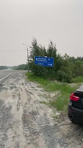 The video shows a roadside scene with a blue directional signpost. The sign points towards Kogalim and Noyabrsk with a distance of 86 kilometers and towards Surgut with a distance of 63 kilometers. The signpost is situated next to a road with a car partially visible on the right side of the frame. The environment appears to be rural with trees and vegetation surrounding the road. The sky is overcast, suggesting cloudy weather. The road surface is paved, with some gravel and tire tracks visible on the side. The video is taken from a stationary perspective.