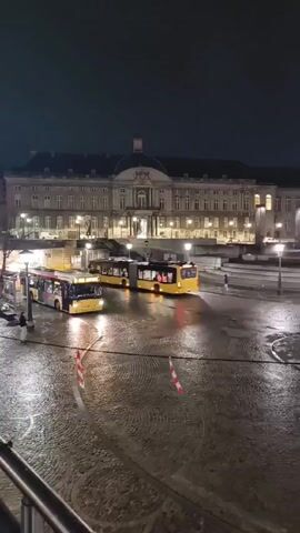 The video shows a night scene of a large building that is illuminated by artificial light. The building has a dome and many windows. In front of the building is a wet, cobblestone plaza, with some caution cones. There are two buses parked in the plaza, as well as several people walking. The sky is dark and overcast.