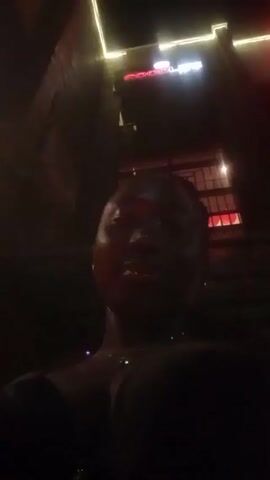 A dimly lit video shows a person's face and upper body. The background features a building facade with a neon sign that reads "GOODLIFE" and a lit window. The person is facing the camera, partially obscured by darkness, making it hard to discern details. The environment seems to be indoors, possibly a bar or lounge, given the lighting and decor.