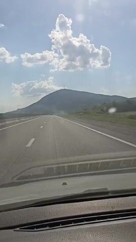 This video shows a view from a car driving on a highway. The car is moving forward on a paved road with white lane markings. To the right of the road, there is a green grassy area and some trees. In the distance, there is a large mountain with a radio tower on top. The sky is blue with fluffy white clouds, and the sun is shining brightly, creating a lens flare in the upper right corner of the frame. The overall scene suggests a sunny day, possibly in the late morning or early afternoon. There is no visible text in the video.
