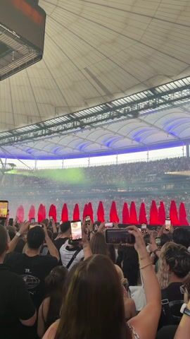 The video opens with a view of a stadium roof and a large crowd of people. The camera then pans down to show a section of the crowd in the foreground, with many people holding up their phones to record. In the middle ground, there are several figures draped in red fabric, appearing to be performers. Behind them, more of the stadium crowd is visible, packed into the stands. The lighting is dim, with spotlights and stage lights illuminating the performers and parts of the stadium. The atmosphere appears to be that of a live concert or performance.