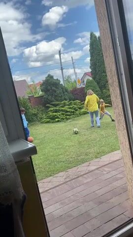 A person in a yellow shirt and blue pants is playing soccer with two children in a grassy yard. The person in the yellow shirt kicks the soccer ball towards one of the children, who is wearing a blue shirt and pants. The other child, wearing a yellow shirt and gray pants, is running towards the ball. The yard is surrounded by trees and a fence, and houses are visible in the background. The sky is blue with white clouds. The video appears to be shot from inside a building, looking out through a window.