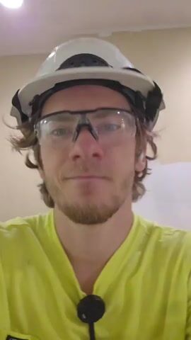 A young man wearing a white hard hat and safety glasses is looking into the camera. He is wearing a bright yellow shirt and has earbuds in. The background is a light-colored wall. He smiles and nods his head.