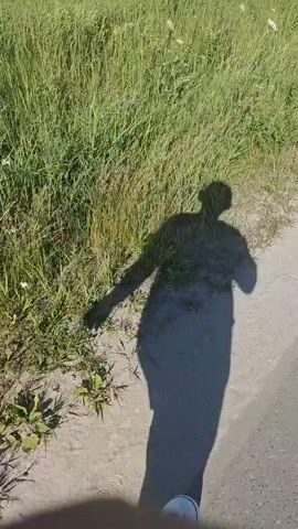 A shadow of a person is cast on a dirt path next to a tall grassy area. The shadow shows the person with arms extended, one arm pointing towards the grass, and the other arm bent at the elbow. The shadow appears to be wearing wide-legged pants. The sun is shining from the right, casting a long shadow to the left. The grass is green and appears to be wild or uncultivated. There are no other people or objects visible in the frame.