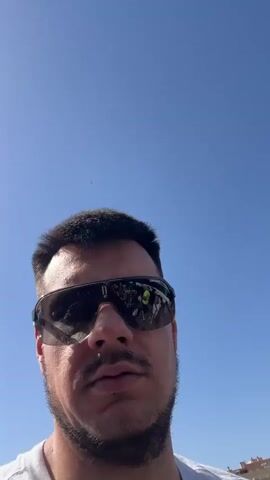 This is a selfie video taken by a man wearing a white t-shirt and large, dark sunglasses with red frames. He has short dark hair and stubble. The video appears to be taken outdoors on a sunny day with a bright blue sky.

The man is standing in what looks like an outdoor market or street fair. In the background, there are rows of stalls set up, displaying clothing and other merchandise. Several people are visible browsing the stalls. There are also buildings in the background, which appear to be apartment buildings, suggesting an urban or semi-urban setting. Palm trees are also visible.

The overall atmosphere seems relaxed and casual, typical of a market day. The bright sunlight and clear sky indicate good weather. The "5€" sign on one of the stalls suggests that prices are low, characteristic of a market. The man's expression is neutral, and he is looking directly at the camera.