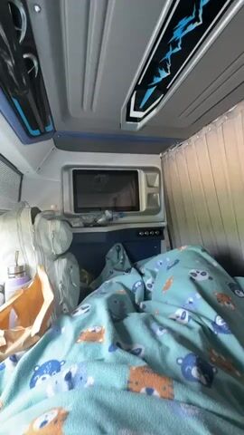 From a first-person perspective, the video shows the interior of what appears to be a traveling sleeper cabin. The camera pans from a small microwave on the wall, above a blue console, towards a window with gray Venetian blinds. On the bed, a light-blue blanket with images of bears and foxes covers the visible area. A white backpack, a water bottle, a purple container, and a brown paper bag sit on the bedside. When the camera focuses on the window, a landscape featuring lush green fields, scattered trees, and a highway with a guardrail becomes visible. The blinds are partially lowered, obscuring part of the view. The interior of the vehicle has a color scheme of blue, gray, and white. There are no people seen in the video.
