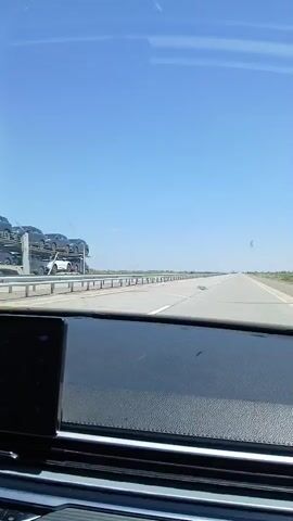 The video shows a view from inside a vehicle as it travels on a highway. The sky is clear and blue with some thin clouds. The highway is paved with multiple lanes and marked with white and yellow lines. A metal guardrail runs along the left side of the road. The landscape on either side of the highway appears to be flat and dry with sparse vegetation. The dashboard of the vehicle is visible in the foreground. The vehicle is moving forward at a steady pace.