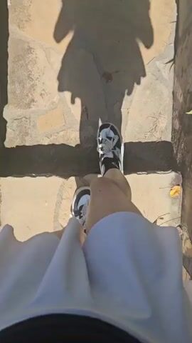 A first-person perspective video shows a person wearing a white skirt and black and white sneakers. The person is walking on a stone path that transitions to a grassy area. The shadow of the person, wearing a skirt, is visible on the ground. The person takes several steps forward, their feet and shadow moving across the path and grass.