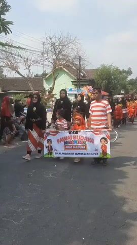 A group of people are walking down a street in a procession. In the foreground, a man in a red and white striped shirt and a woman in traditional Indonesian clothing are holding a banner that reads "PAWAI BUDAYA NUSANTARA ADAT MADURA" and "TK AL HIDAH JAMBEWANGI 03". Several children in traditional Indonesian attire are walking behind them. In the background, more people are walking in the procession, some of whom are carrying balloons. The street is paved and lined with houses. The sky is bright and sunny.