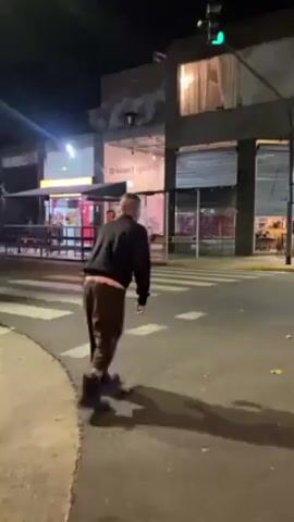The video shows a person performing a handstand in the middle of a crosswalk at night. They are wearing dark clothing with light-colored pants visible underneath their jacket. The crosswalk is located on a street corner with buildings in the background. A traffic light is visible and appears to be green. The person holds the handstand briefly before returning to an upright position. The scene is illuminated by streetlights and lights from nearby businesses. The video appears to be taken from a stationary position.
