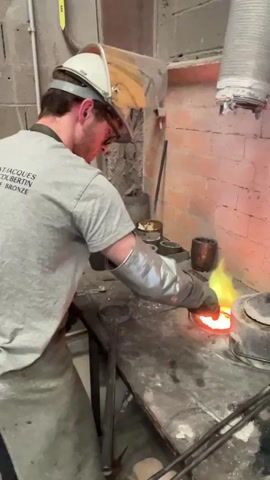In a workshop or foundry setting, a man wearing a protective helmet with a face shield, a grey t-shirt, an apron, and heat-resistant gloves is actively engaged in metalwork. The grey t-shirt has visible text that reads "JACQUES DOUBERTIN BRONZE".

The man is using a metal tool to stir a crucible filled with intensely glowing, molten metal. Bright orange and yellow molten material is visible within the crucible, emitting vibrant green and yellow flames or smoke that rise above the surface. The crucible rests on a sturdy workbench alongside other metal containers and tools, suggesting an industrial or artisanal metalworking environment. A brick wall forms the background, and a ventilation duct is visible overhead. The scene captures a moment of precise and careful handling of extremely hot material in the process of metal casting or melting.