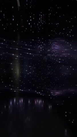 The video shows a dark room with numerous small, bright lights scattered across the ceiling and walls, resembling a starry sky or a galaxy. Some of the lights appear as single points, while others are elongated streaks, possibly indicating movement or a long exposure effect. There are also faint reflections of these lights on a dark, glossy floor. The overall impression is of a futuristic or immersive environment, possibly a planetarium or an art installation. There are no people or discernible objects in the scene. The lighting is primarily dark with accents of purple and white.