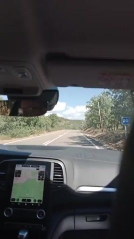 The video shows the interior of a car from the perspective of a passenger. The driver's head is visible in the rearview mirror. A navigation screen is displayed on the dashboard, showing a map. The car is driving on a paved road through a rural area with trees on either side. The sky is blue with some clouds. A speed limit sign is visible on the right side of the road.