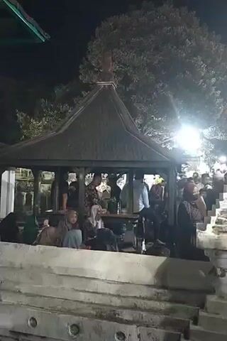 The video shows an outdoor scene at night. A group of people are gathered around a covered structure with a pyramid-shaped roof. Some individuals are seated within the structure, while others are standing. There are also people visible on a set of stairs to the right of the structure, and further back in the background. A large tree with foliage is visible above the structure. Several lights illuminate the area, casting shadows. The overall atmosphere appears to be casual, with individuals interacting amongst themselves. The ground surface is rough and appears to be made of stone or concrete.