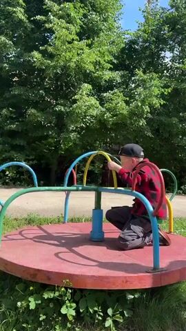 The video shows a young boy on a playground merry-go-round. He is wearing a red and black checkered shirt, dark pants, a black baseball cap with a Minecraft logo, and glasses. The merry-go-round is circular with colorful metal poles and arches in red, blue, and yellow. The boy is holding onto the bars and appears to be enjoying himself, with an excited expression. He is positioned in the center of the merry-go-round, which is red.

The setting is outdoors, with lush green trees and foliage surrounding the playground. The ground is paved, and there is some grass around the base of the merry-go-round. The sunlight suggests it is daytime, likely a sunny day given the bright lighting and shadows. A building with a window and a green sign with a number "6" is visible in the background. The overall impression is a typical, cheerful scene of a child playing at a local park in Yunost', Russia.