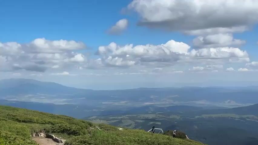 The video shows a panoramic view from a mountain summit. In the foreground, there is lush green vegetation and some rocky terrain. A dirt path is visible on the left side. Further back, a railing with some people standing near it is seen on the slope. The background reveals a vast expanse of rolling green mountains and valleys fading into the distance under a bright blue sky with scattered white clouds. The overall impression is one of a serene and scenic mountainous landscape on a clear day.