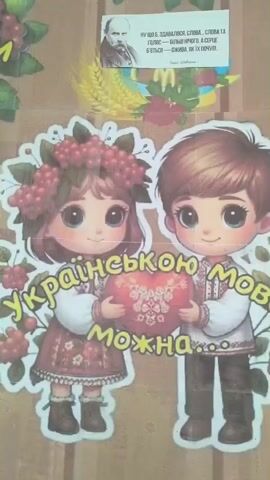 The video shows a colorful poster or wall decoration featuring two cartoon children in traditional Ukrainian clothing holding a heart. The girl on the left wears a floral wreath and a red embroidered dress, while the boy on the right wears a white embroidered shirt and brown pants. The text on the poster reads "Українською мовою можна" which translates to "In Ukrainian language, it is possible." Below the children, in separate heart shapes, are the words "писати" (to write) and "говорити" (to speak). In the upper right corner of the poster, there is a smaller image of a man, likely a historical figure, with some Ukrainian text above him. The background of the poster appears to be a wooden or textured surface. The overall impression is a patriotic or educational display celebrating the Ukrainian language.