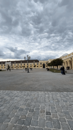 Tourists visit Schönbrunn Palace courtyard under cloudy Vienna skies