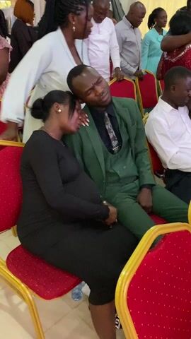 The video depicts a group of people seated on chairs in what appears to be an indoor setting. The chairs are red with gold accents. At the beginning of the video, a pregnant woman in a black dress is seated next to a man in a green suit. The woman has her head resting on the man's shoulder. The camera then shifts focus, showing other individuals in the room, some engaged in conversation, others looking around. The camera movement is shaky, and the focus shifts quickly, making it difficult to discern details about all the individuals present. The environment seems to be well-lit with light coming from unseen sources. The video ends with a quick pan across the room, showing more attendees seated on chairs.