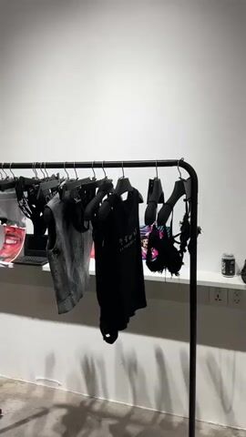 The video shows a group of people in what appears to be a fashion event or retail space. In the foreground, a clothing rack displays several black garments, including a vest and a dress. A woman in the background is taking a photo with her phone, and another person is reflected in a mirror. Further back, more people are gathered, some are holding drinks, and others are looking at their phones. The environment is well-lit with white walls and a ceiling with track lighting. The overall atmosphere seems social and perhaps a bit crowded.