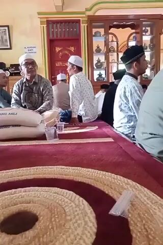 A group of men are gathered indoors, seated on a carpeted floor. The room appears to be a prayer hall or a similar religious space, with ornate wooden carvings and decorated walls. Several men are wearing traditional Islamic attire, including kufis and long robes. In the foreground, a man in a light green shirt and dark patterned pants is looking down. To his right, a man in a maroon shirt and sarong is speaking. Between them are several large sacks, possibly containing rice, and small plastic cups. Other men are visible in the background, some in white shirts with patterned designs, and one wearing glasses. The lighting suggests it is daytime, with natural light coming from unseen windows. The overall atmosphere seems to be one of a gathering or a religious ceremony. The audio consists of Islamic chanting or recitation.