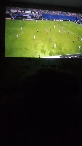 This media file captures a screenshot of a soccer match being displayed on a television screen. The scene depicts a bright green soccer field with several players in white and dark uniforms scattered across it. White lines mark the boundaries of the field, including a penalty area and a goal. In the lower left corner of the screen, a scoreboard shows the time as 00:57 and the score as 0-0. Text labels like "ENG" and "ARG" suggest the match is between England and Argentina. There are also smaller text elements that are difficult to read, but "FIFA" is discernible. The lighting suggests it could be daytime or a well-lit stadium, and the field appears to be in good condition. The right side of the screen is dark, likely due to the camera's angle or the room's lighting, obscuring part of the television image. The overall impression is of a paused moment in a professional soccer game.